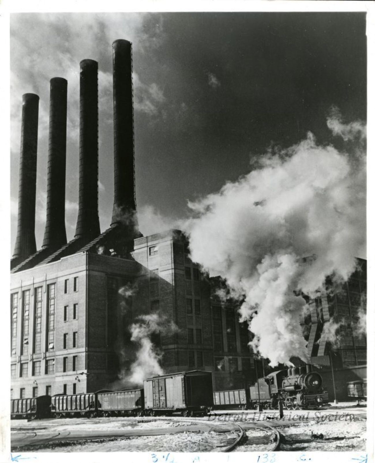 Black and white photo showing the Detroit Edison Delray electric power plant. The view shows railroad tracks, a boxcar, and coal hopper cars in the foreground along with New York Central Railroad locomotive #8558.