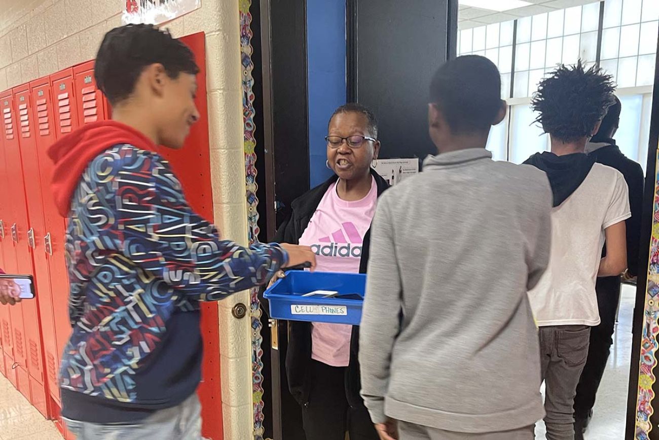 a teacher holding a blue bin and kids putting their cellphones in there