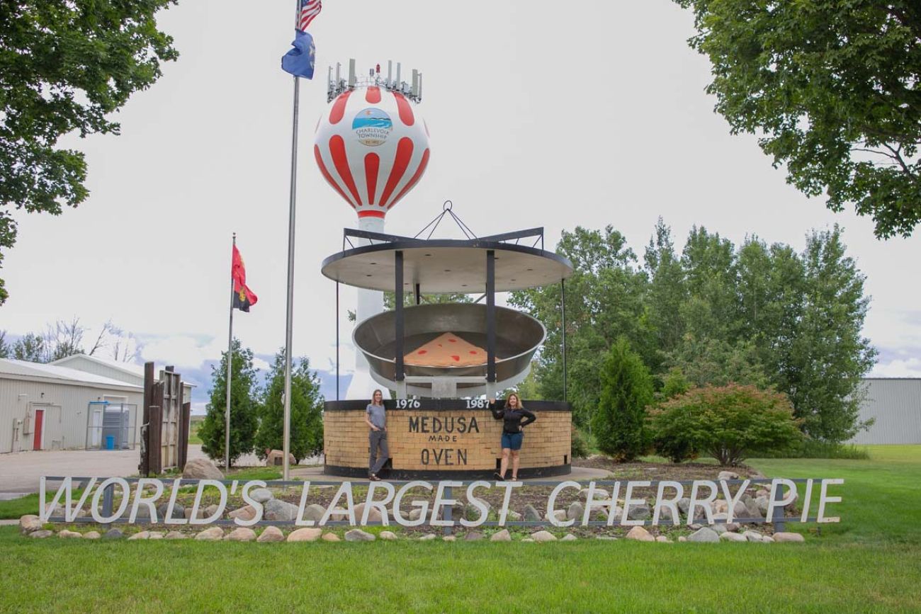 A statue of the "world's biggest pie." Two reporters stand next to it