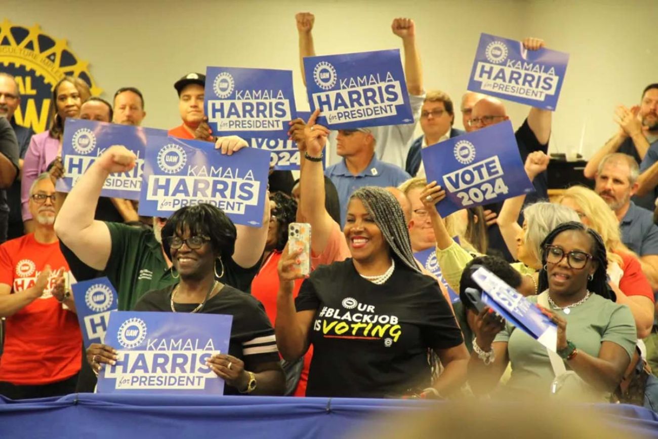 A crowd blue "Kamala Harris for President" signs with an UAW logo