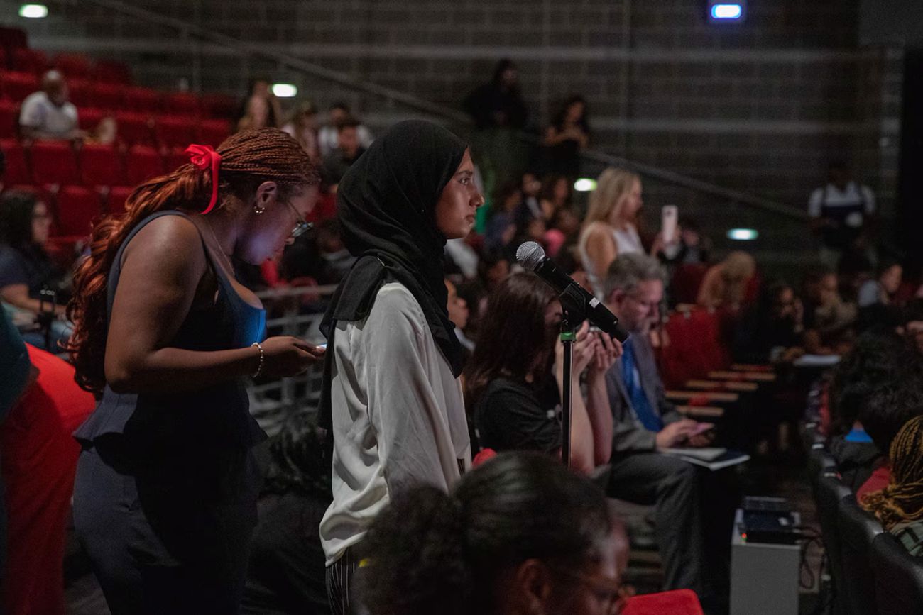 Students sitting in auditorium in Detroit. One is standing to speak at a microphone