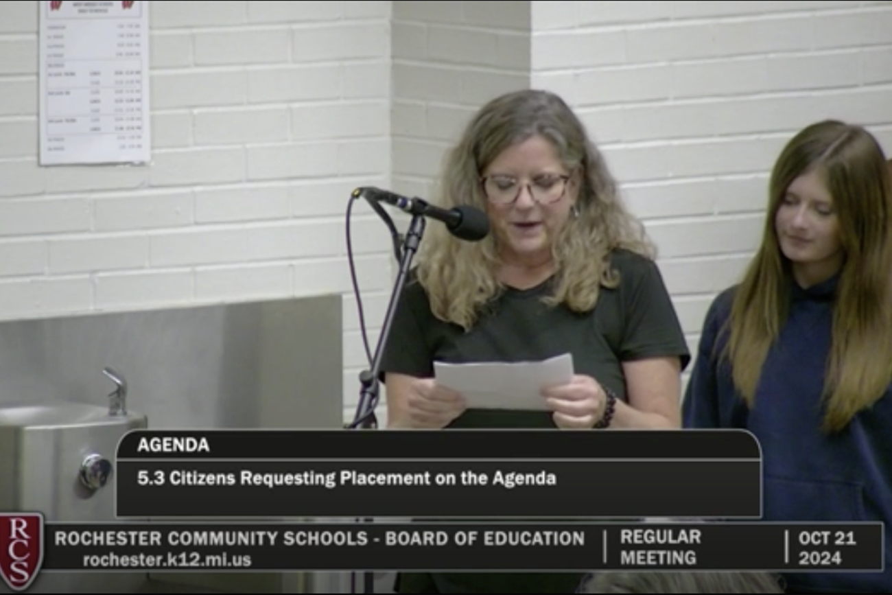 A screenshot of Kristi Fleming speaking into a microphone at school board meeting for Rochester Community Schools