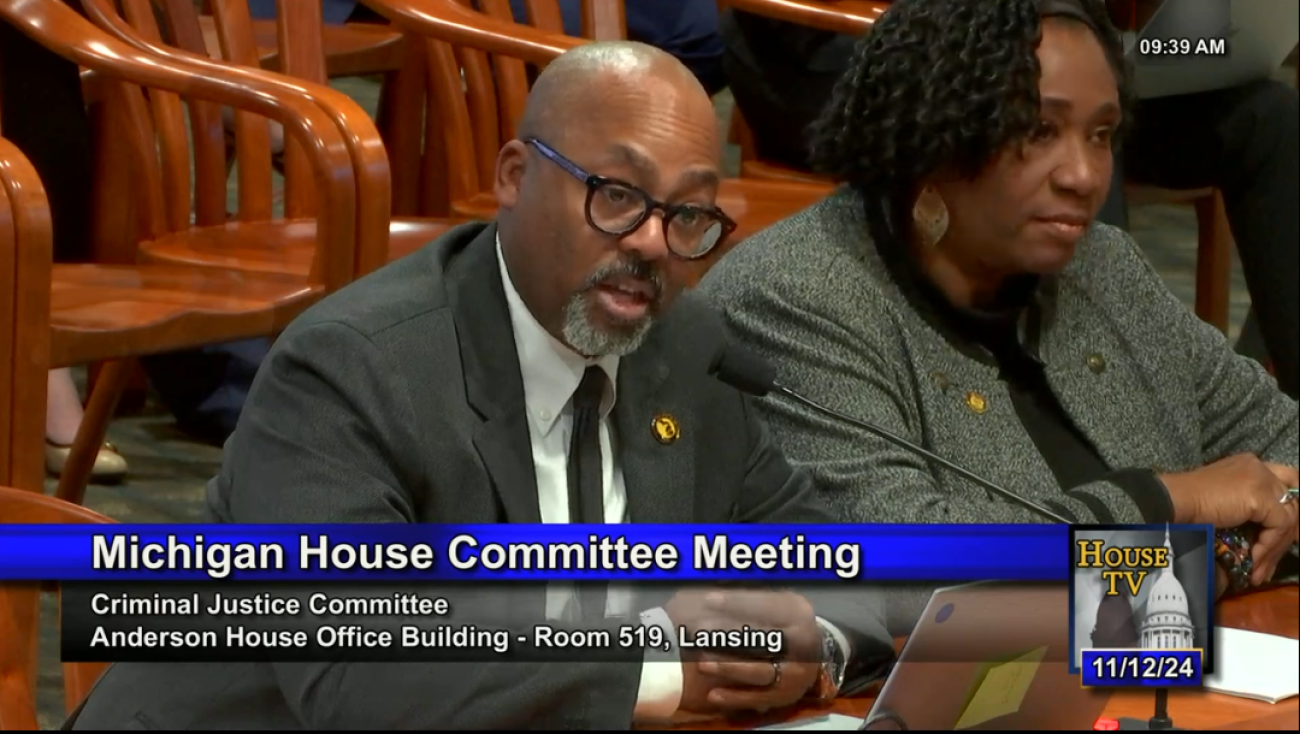 William McConico is sitting at a table, speaking into a microphone