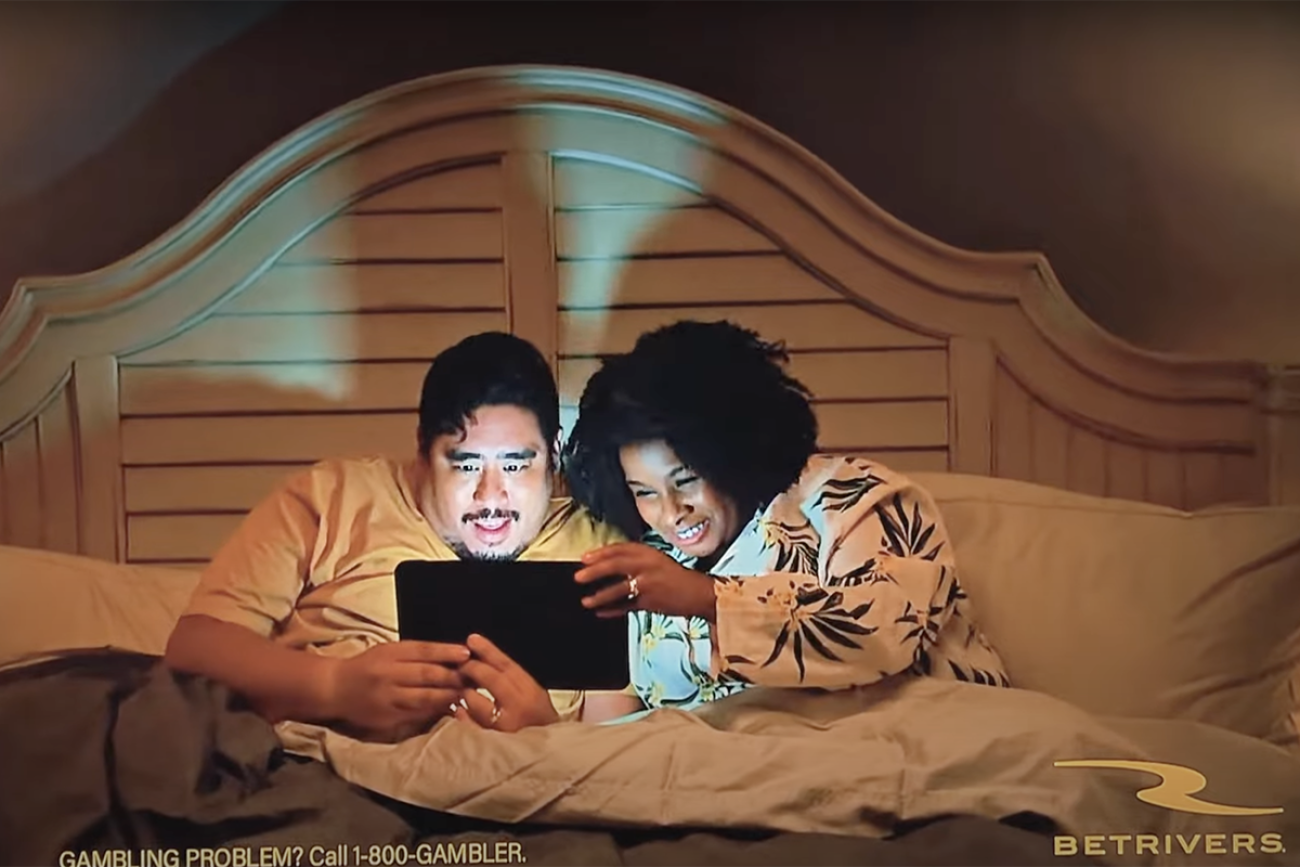 A couple in bed looking at a tablet in an ad for BetRivers