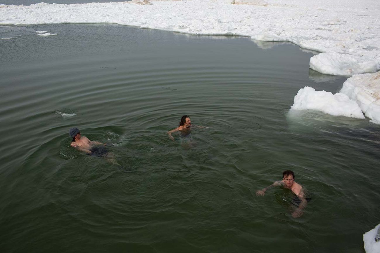 Three people swimming in water. There's snow on the ground. 