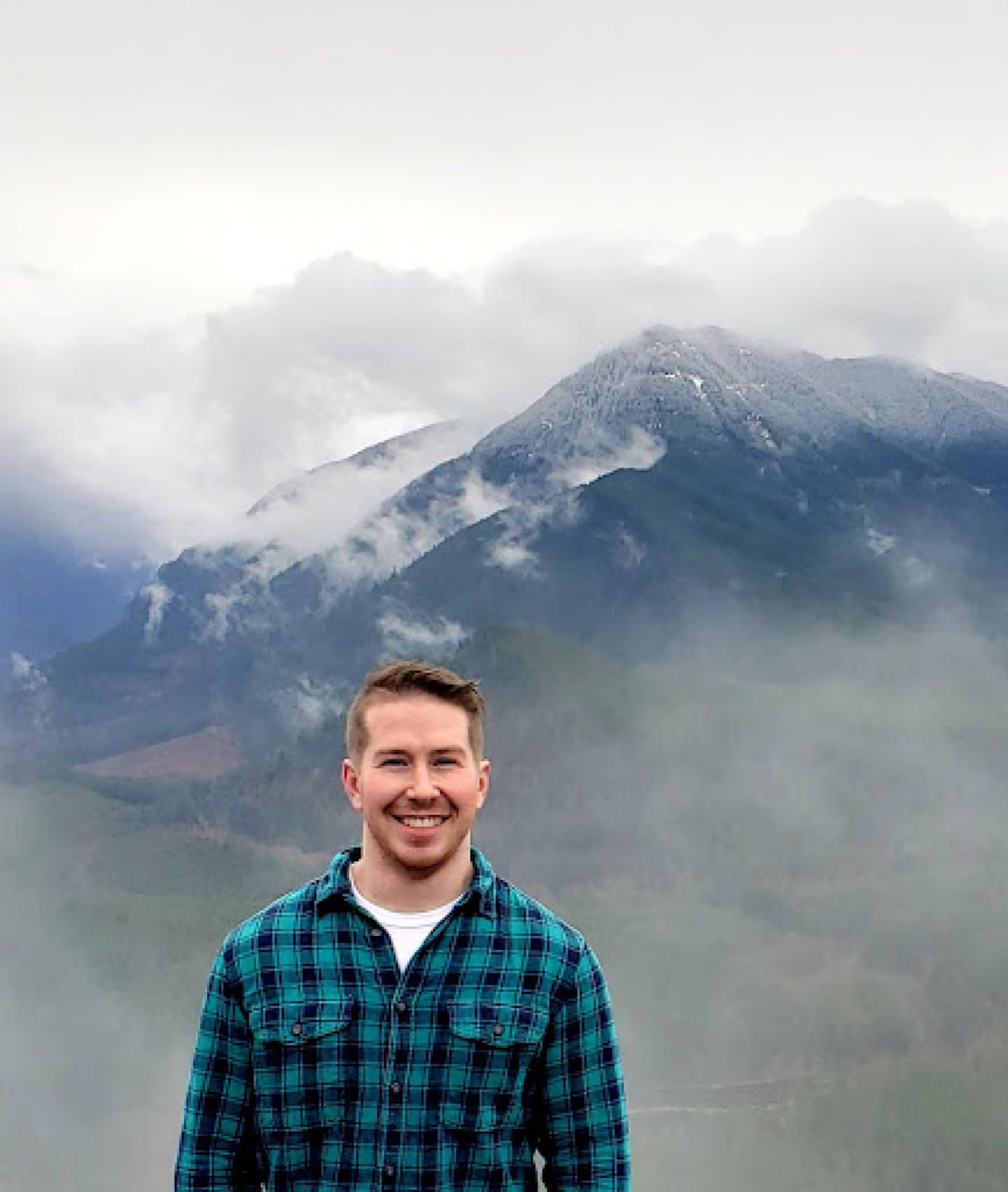 Tim Minotas is wearing a flannel shirt in front of a mountain. 