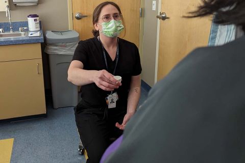 A woman in dark-colored scrubs and a face masks holding a cup of pills 