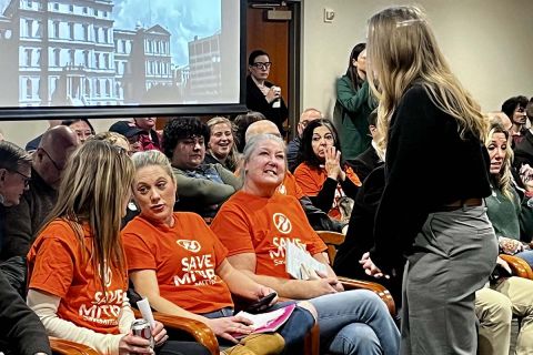 A group of people, many wearing orange t-shirts that says "Save MI Tips" at a committee hearing 