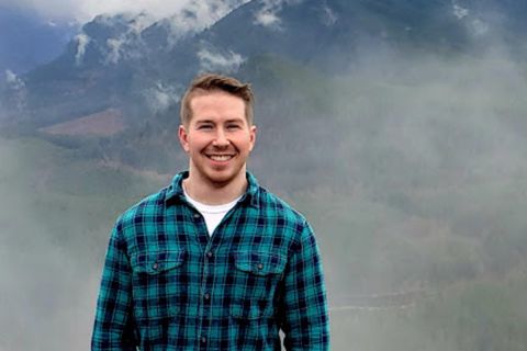 Tim Minotas is wearing a flannel shirt in front of a mountain. 